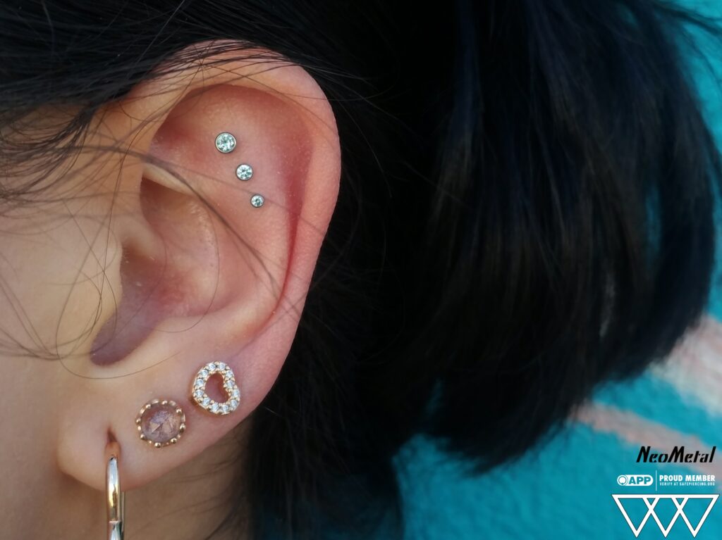 A triple flat piercing featuring bezel-set Swarovski gem ends from NeoMetal, showcasing precise alignment and varying gem sizes that add dimension to the ear's upper cartilage.