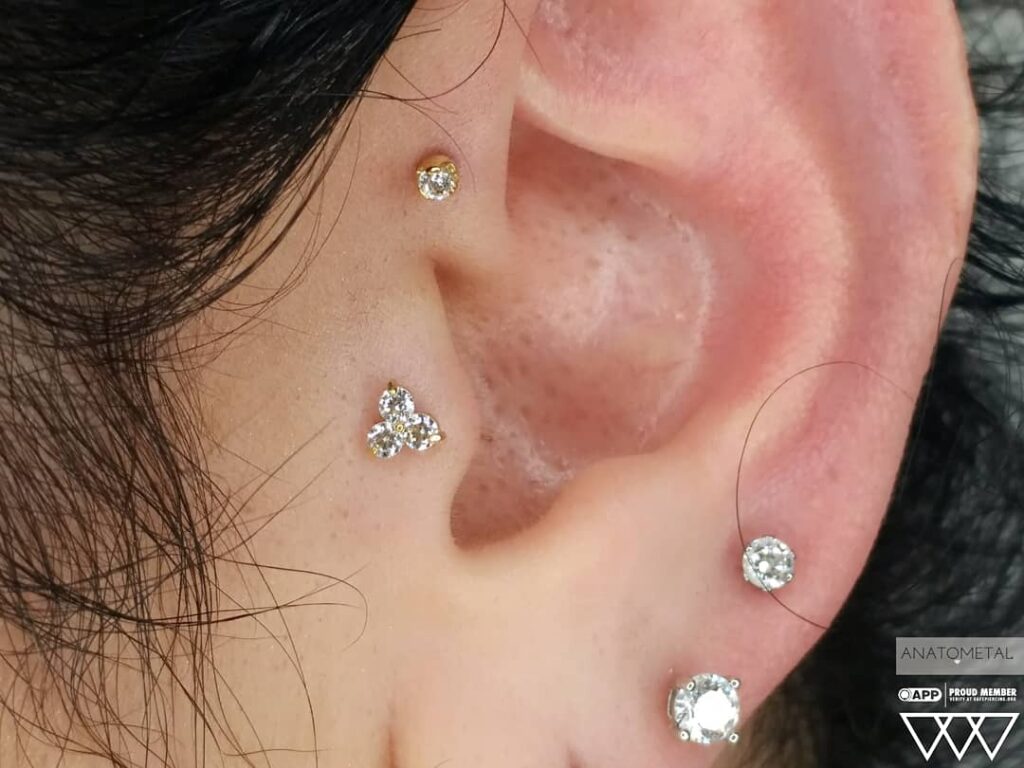 Close-up of a tragus piercing featuring an 18k gold “Trio” end from Anatometal, composed of three clustered round gemstones. The polished gold and symmetrical design add a refined sparkle to the ear's cartilage flap.
