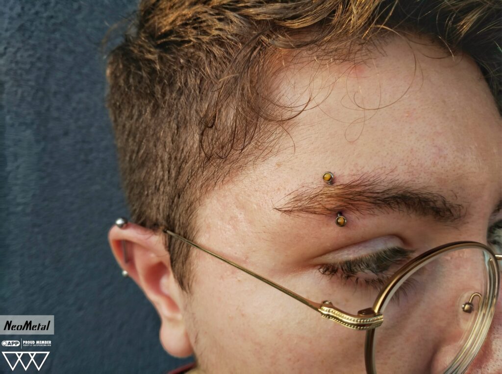 Vertical eyebrow piercing featuring a NeoMetal implant-grade titanium curved barbell, bezel-set with a tiger’s eye gemstone. The warm, golden-brown stone adds a subtle yet striking accent above the outer brow.