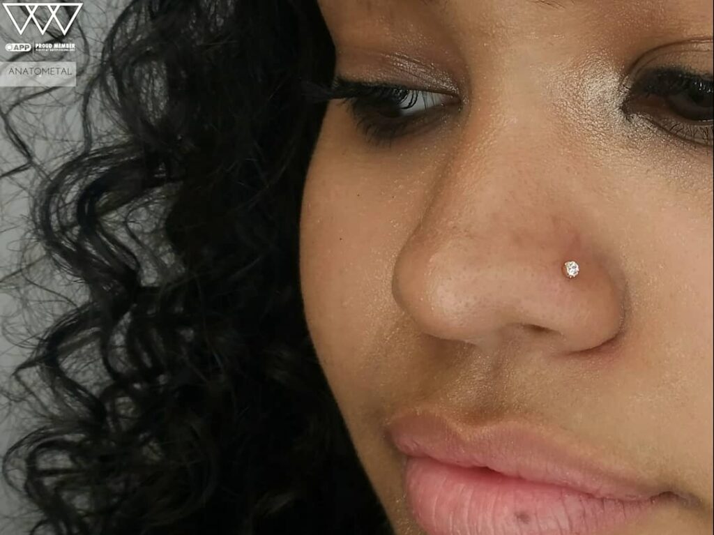 Close-up of a nostril piercing featuring an 18k yellow gold prong-set gemstone from Anatometal, positioned on the left nostril. The polished gold setting and sparkling gem add a subtle yet elegant touch to the wearer's profile