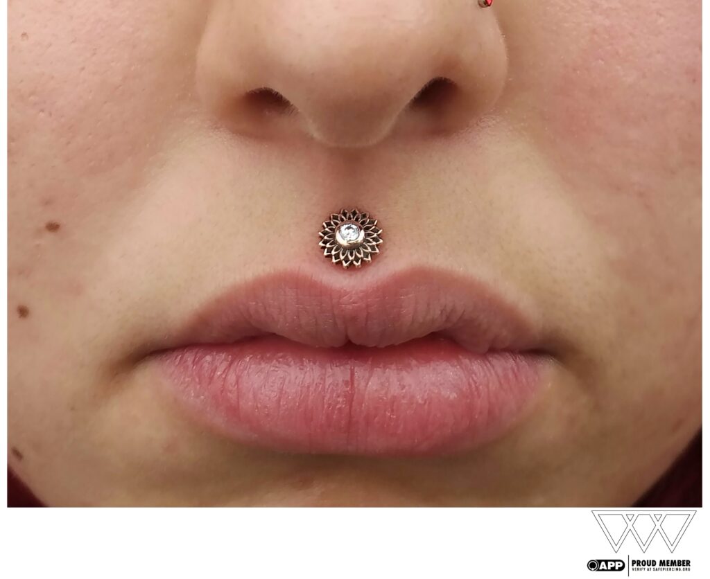 Close-up of a philtrum piercing featuring a 14k gold “Thrive” end from Maya Jewelry, positioned just above the upper lip. The textured design and polished finish add a bold yet elegant touch to the wearer's facial features.