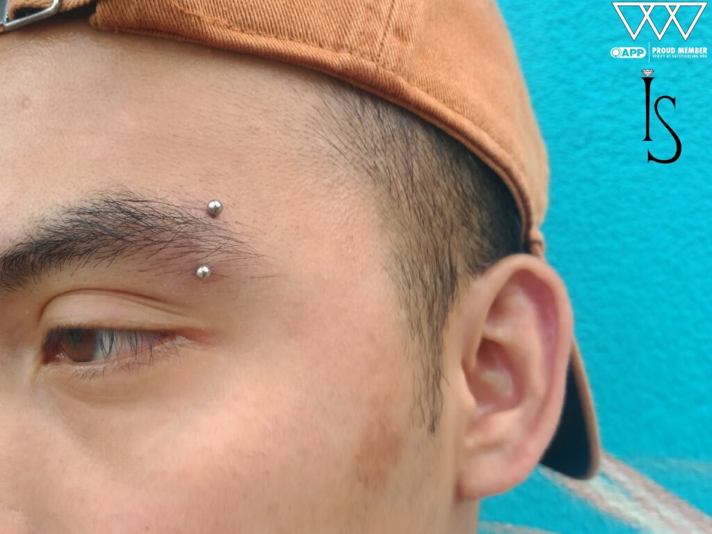 Close-up of a vertical eyebrow piercing featuring a titanium curved barbell from Industrial Strength. The jewelry sits neatly along the brow ridge, with its polished finish catching the light against the skin.