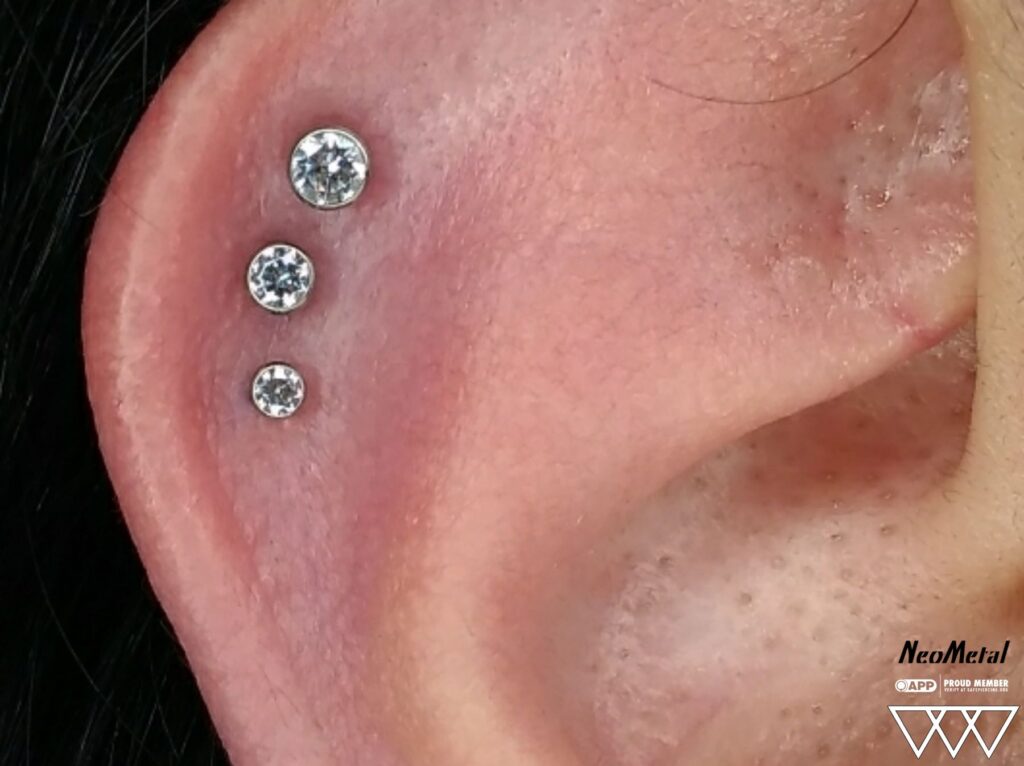 Close-up of a triple helix piercing featuring three graduated bezel-set titanium ends with Swarovski gemstones, aligned vertically along the ear’s outer cartilage.